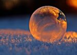 Close-up shot of a frozen bubble with warm reflections resting on a snowy surface at twilight.